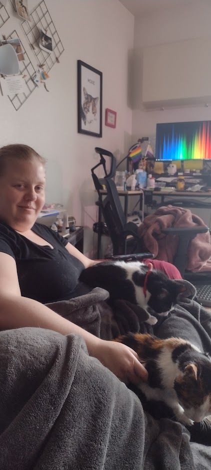 Ivory smiles at the camera with the tuxedo cat Kochanski and the calico cat Moxie rest in her lap. They may not be co-existing peacefully in this moment.