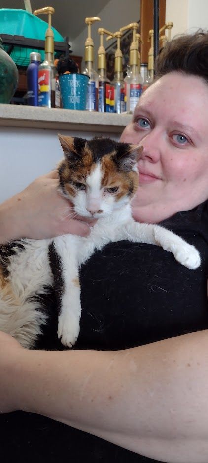 Kate and Moxie the calico cat pose together for the camera. This is their last photo together. Kate has dark brown hair and blue eyes, they are wearing a black shirt covered in cat hair.