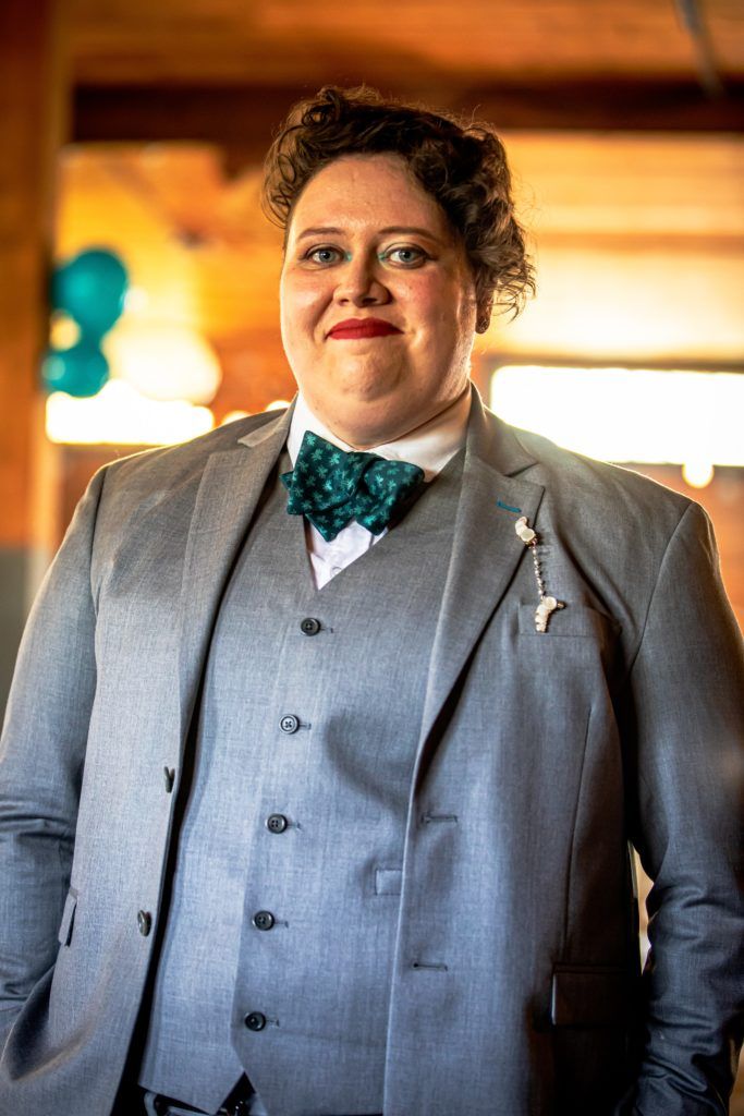 Kate smiles at the camera on their wedding day, wearing a gray suit and teal bow tie. The tie has small light teal octopuses on it.