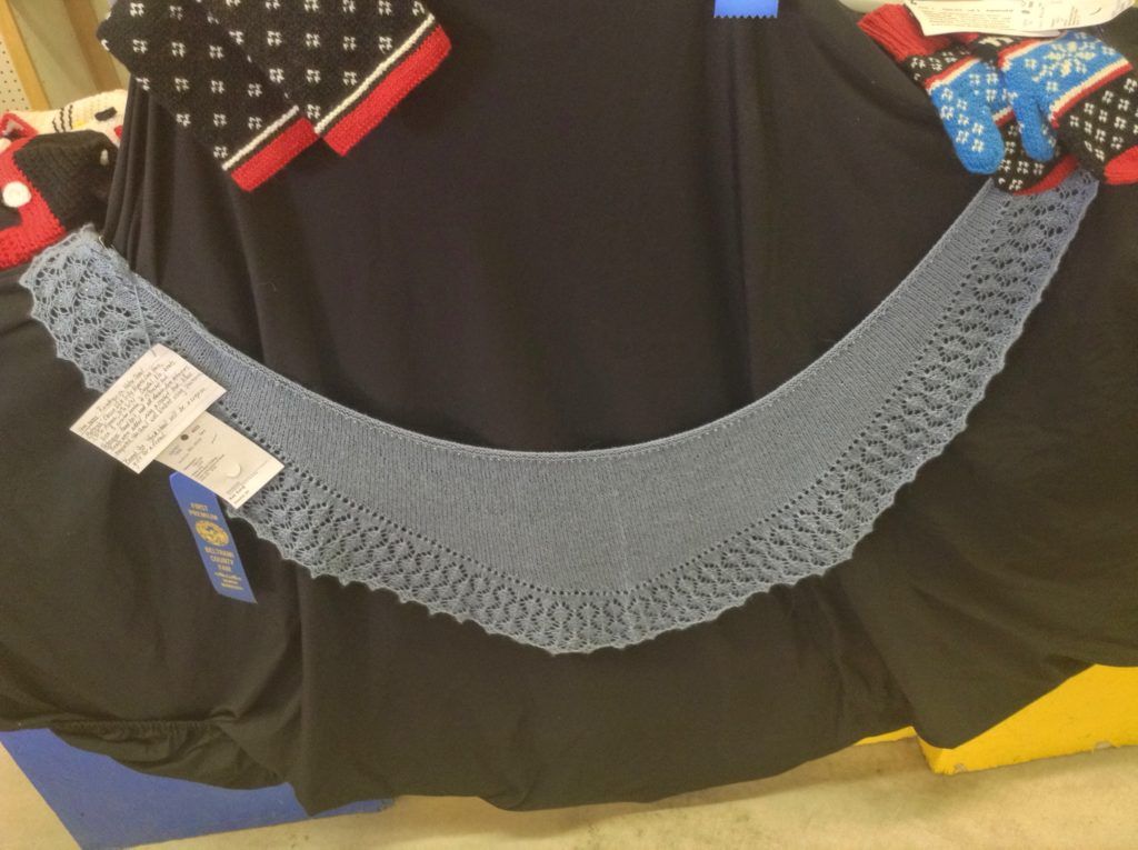 the light blue shawl hangs on a black background with a blue fair ribbon pinned to it