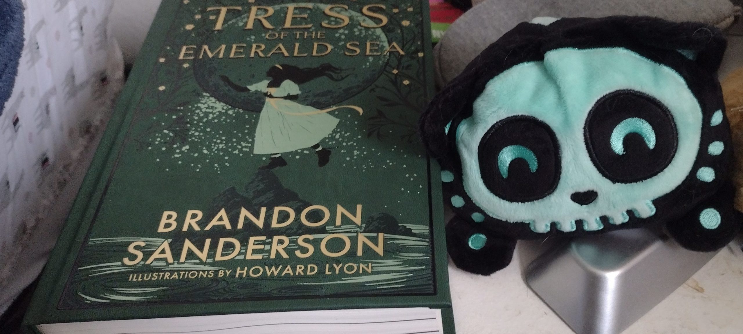 A foiled copy of Tress of the Emerald Sea sits on a nightstand next to the skeleton stuffy of a Soonie Pup.