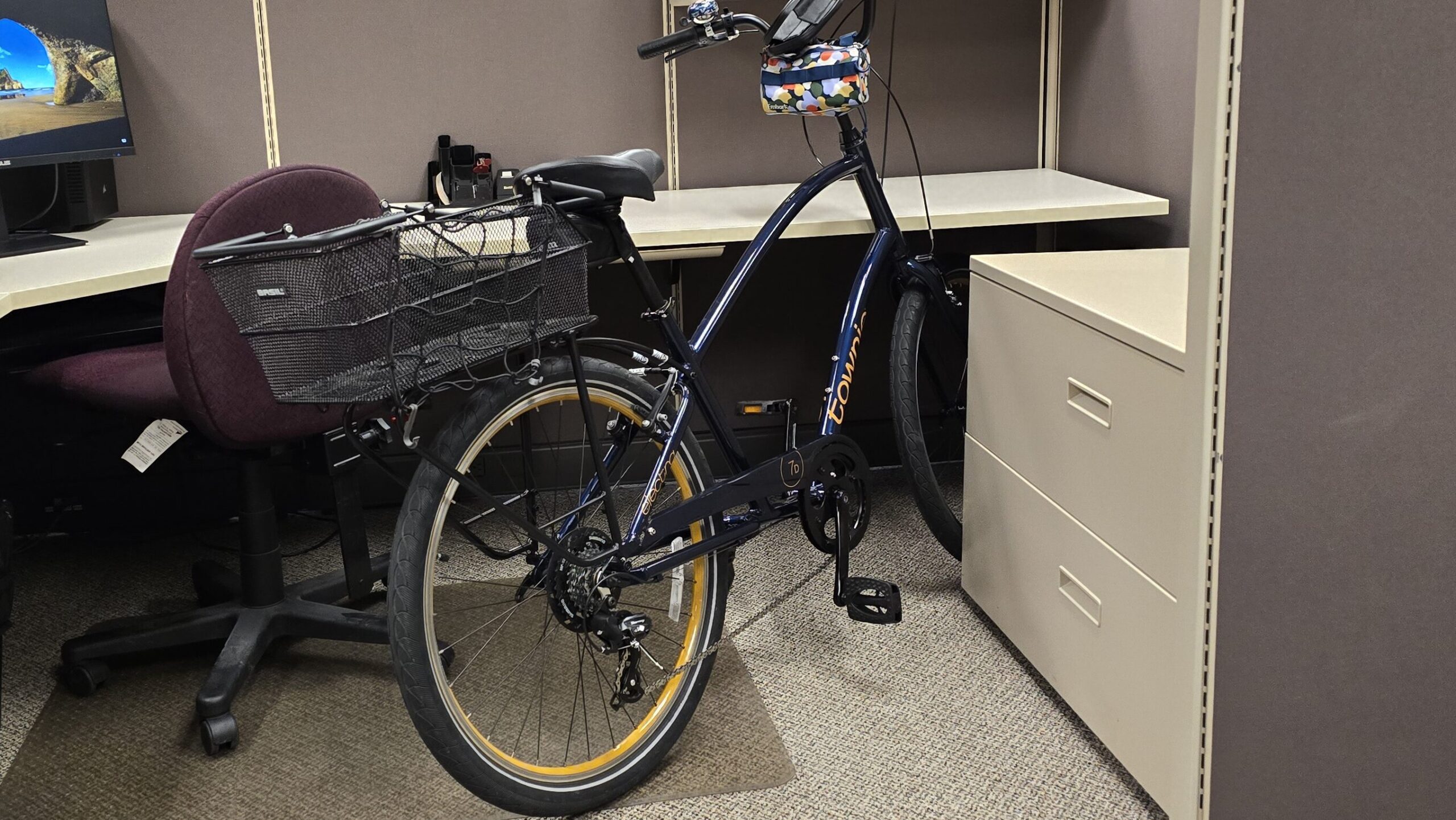 A dark blue Electra Townie 7D step-over bike with a rear rack and basket and a floral camouflage handlebar bag sits in on otherwise empty office cubicle.